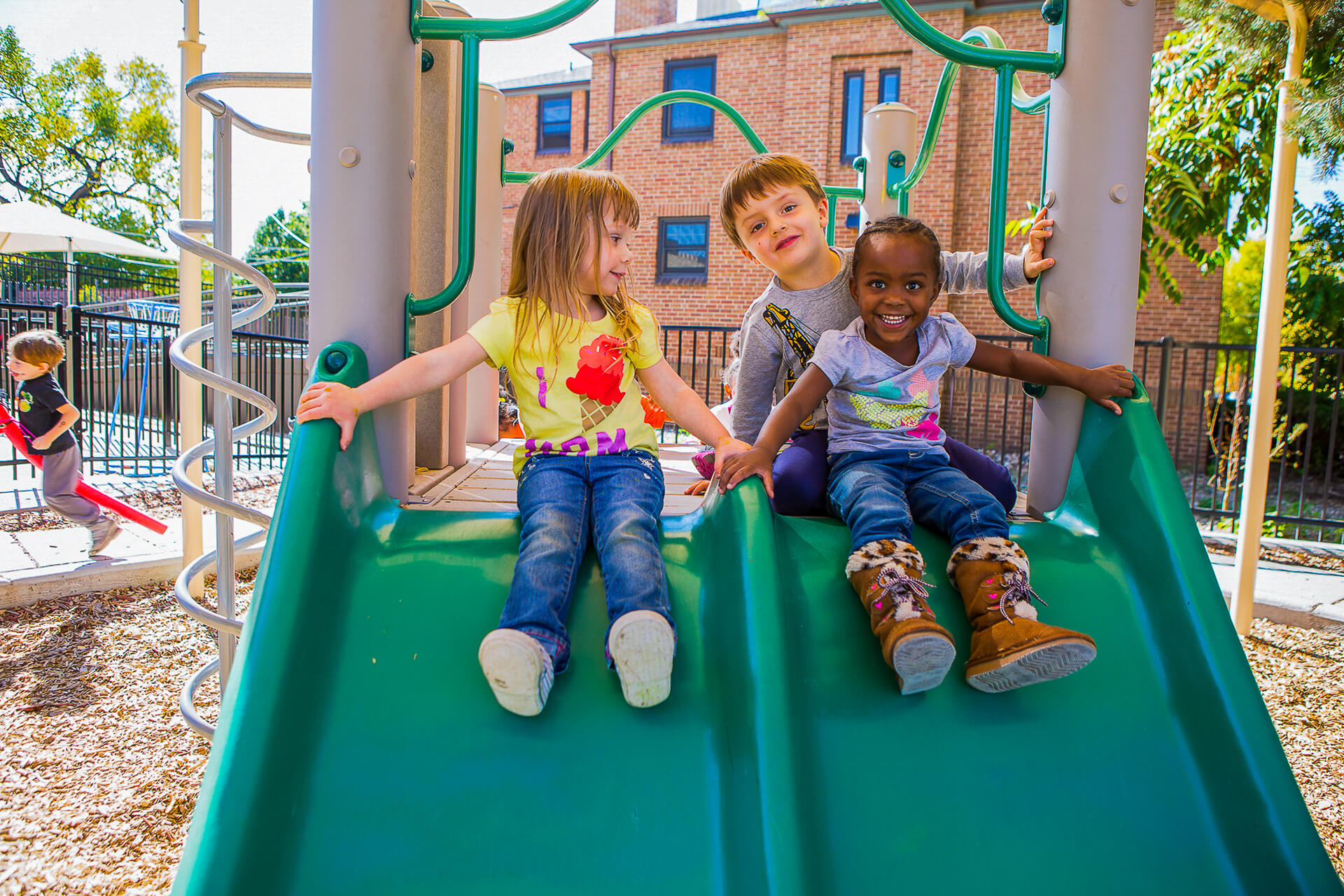 Three diverse children smile at the top of a green playground slide. A brick building and trees visible in the background.