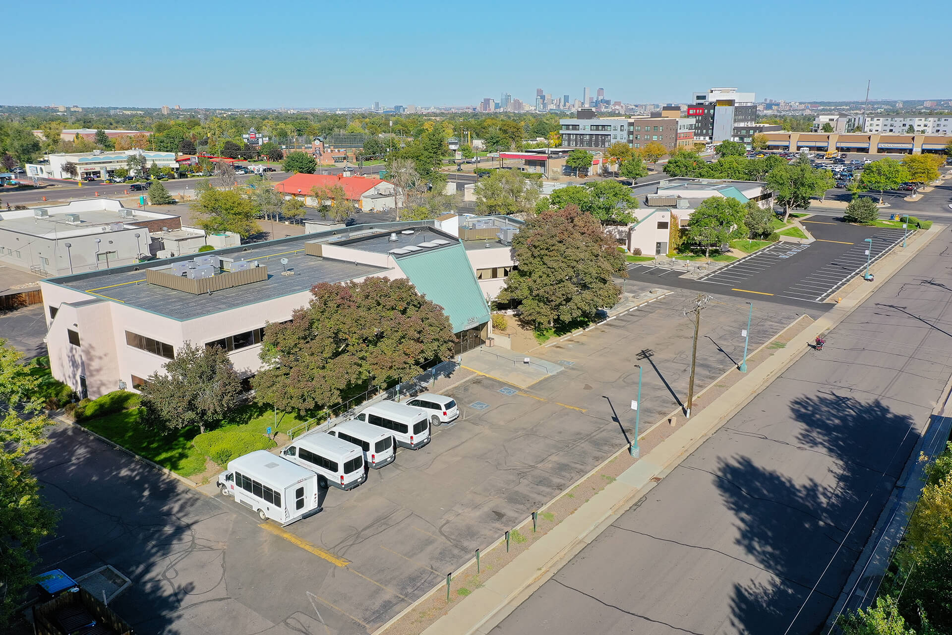 Aerial view of a white and green building in a city surrounded by trees. Five white vans are parked in the parking lot.