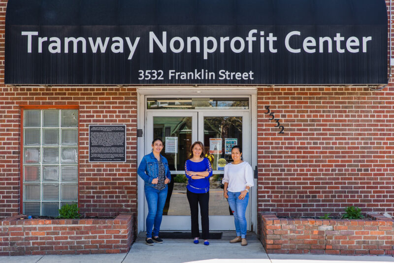 Three people in front of brick building with sign: Tramway Nonprofit Center