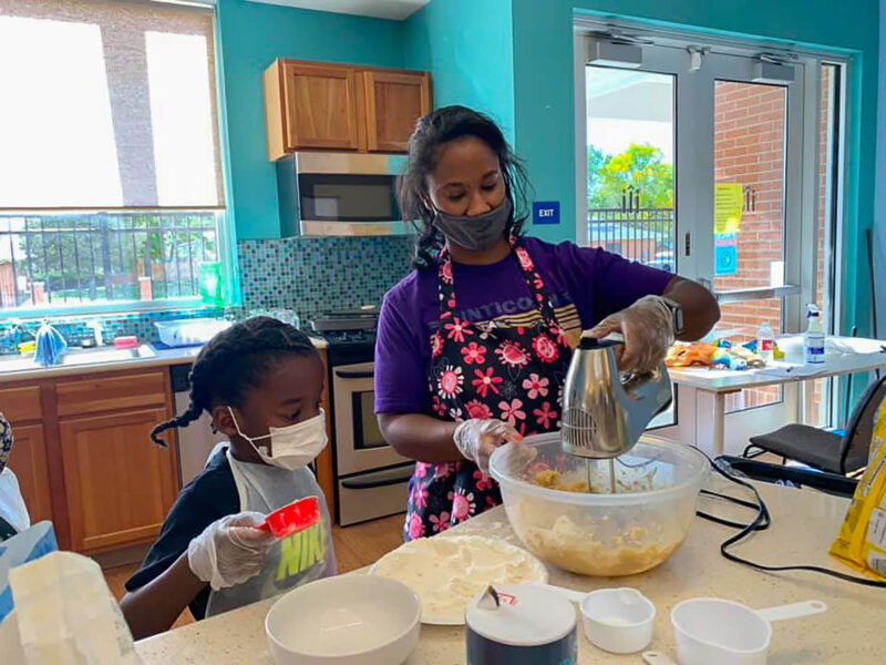 Young girl watching woman mix in a bowl, in a teal kitchen