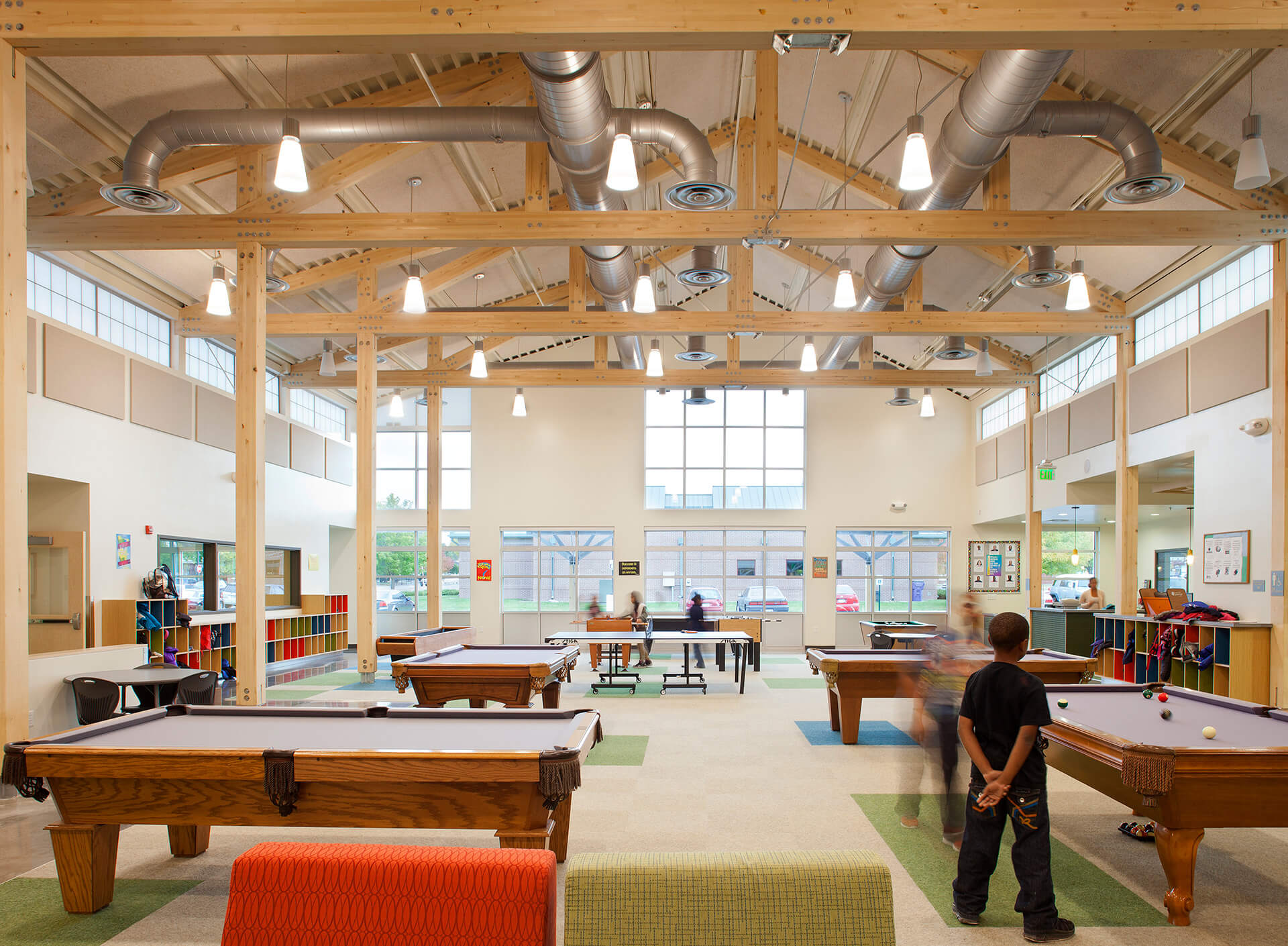 A spacious recreation room featuring wooden beams, exposed ductwork, and large windows. Several pool tables and a ping pong table are scattered throughout the room. Children are actively playing games while others are socializing. The space is bright and modern.