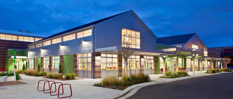 A modern community center building photographed at twilight, featuring large windows illuminated from within. The exterior includes clean architectural lines, metal siding, and a covered entryway with decorative beams. A row of bike racks and landscaped greenery lines the sidewalk.