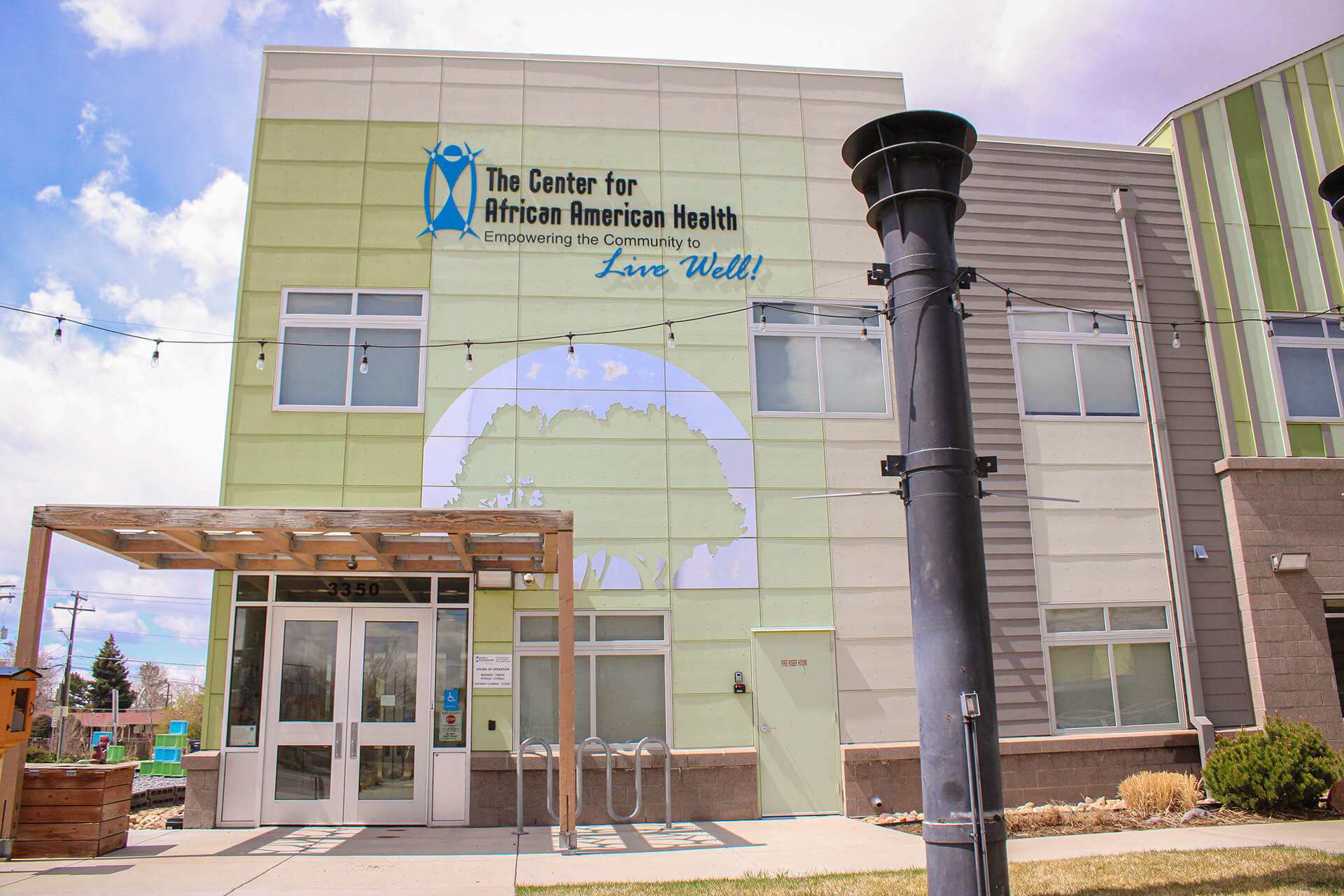 The Center for African American Health building exterior, painted in shades of green and beige, with the organization's logo and mission statement displayed prominently on the facade. A wooden pergola structure sits above the main entrance, and a tall black chimney-like structure is in the foreground.