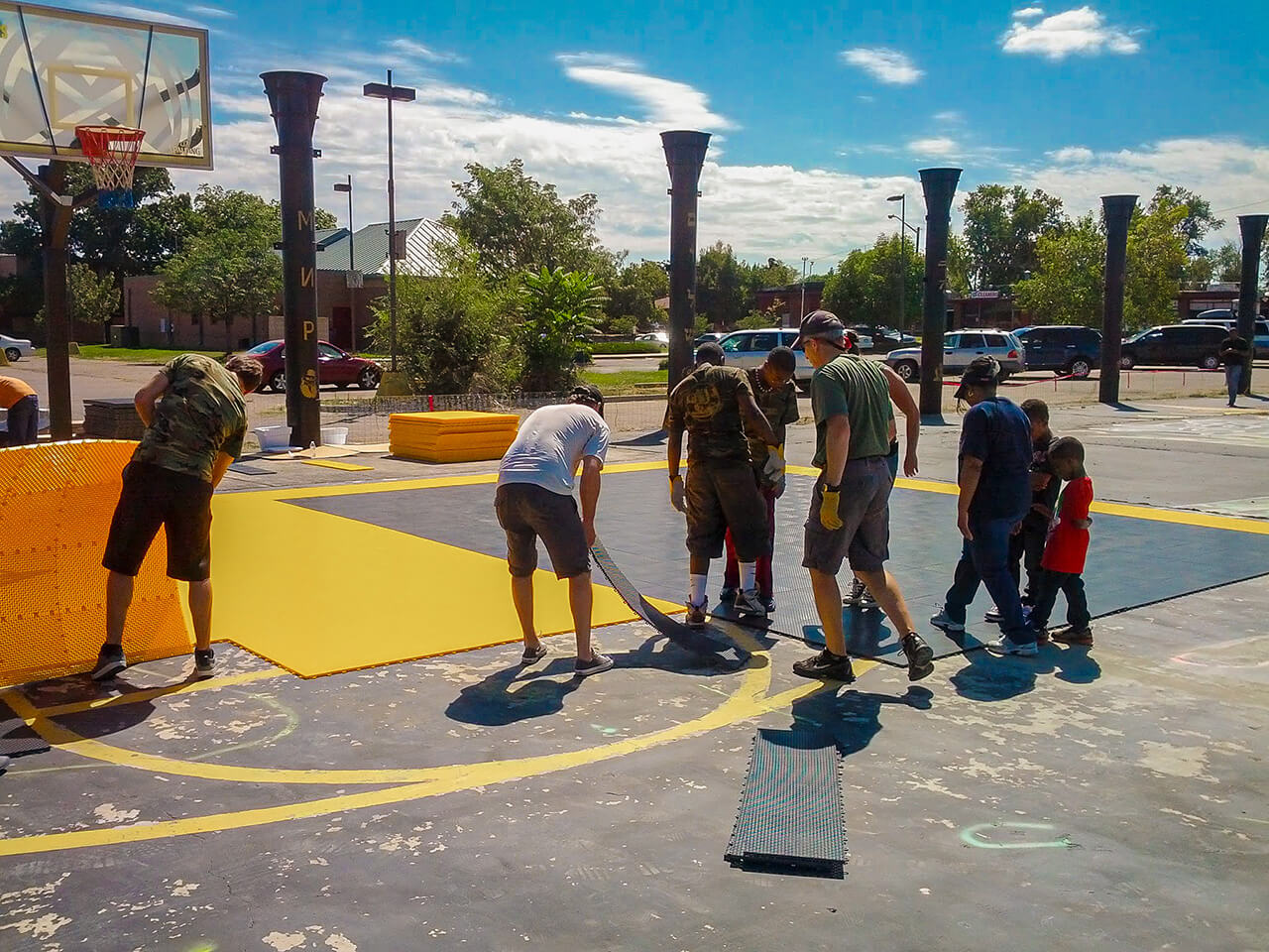 A group of volunteers working outdoors to construct or refurbish a basketball court. They are installing bright yellow and black flooring panels under a sunny sky. Nearby, basketball hoops, parked cars, and trees are visible in the background.