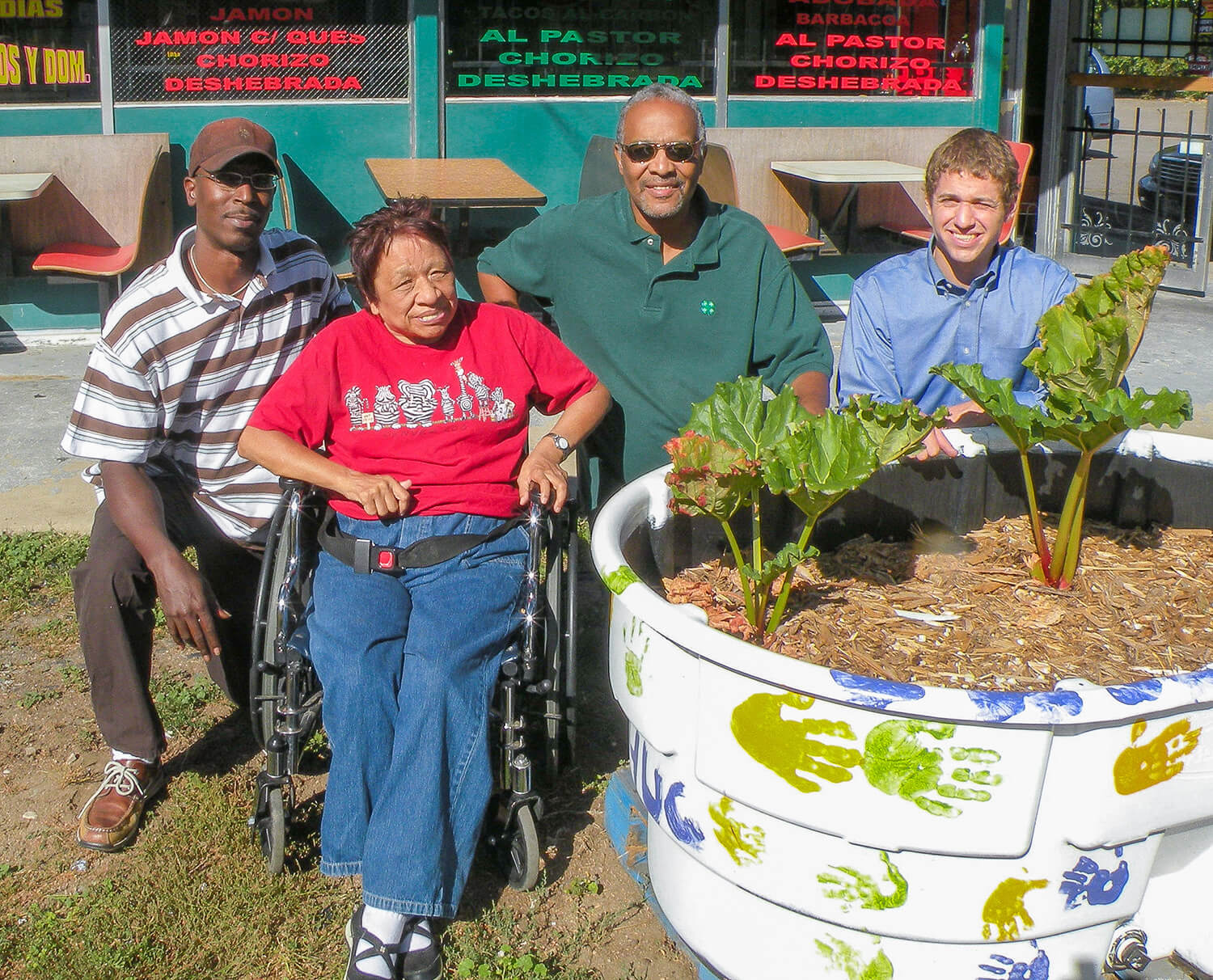 Four individuals posing next to a community garden planter painted with colorful handprints. The planter contains green plants growing in soil. One person is seated in a wheelchair, and the others stand nearby, smiling. A green building with red signage is in the background.