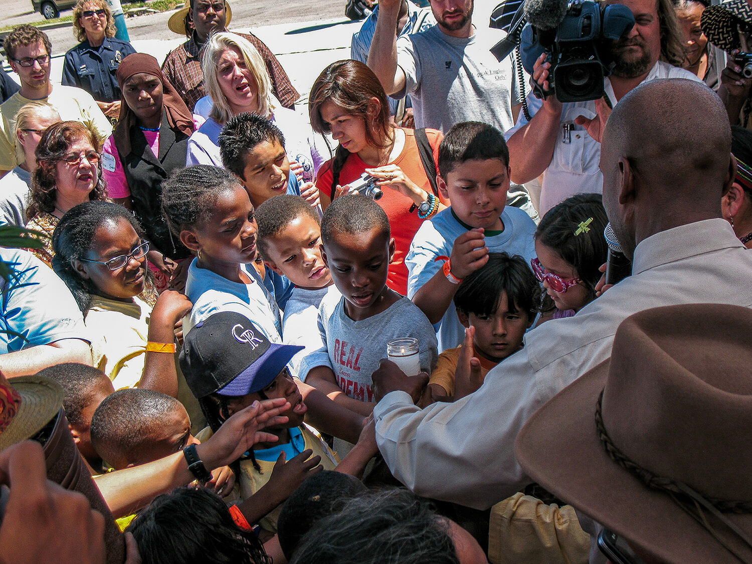 A diverse group of children and adults gathered around a man in a white shirt holding a candle at a community event.