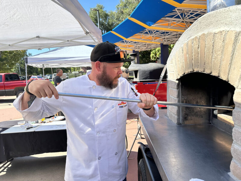 Man holding a pizza peel in a large pizza oven, set outside