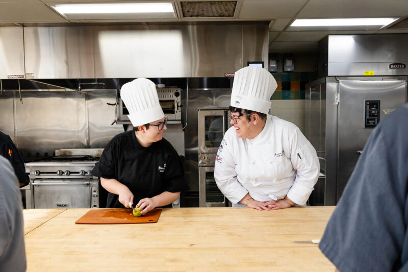 Two female chefs wearing chef hats cutting on a large wooden table