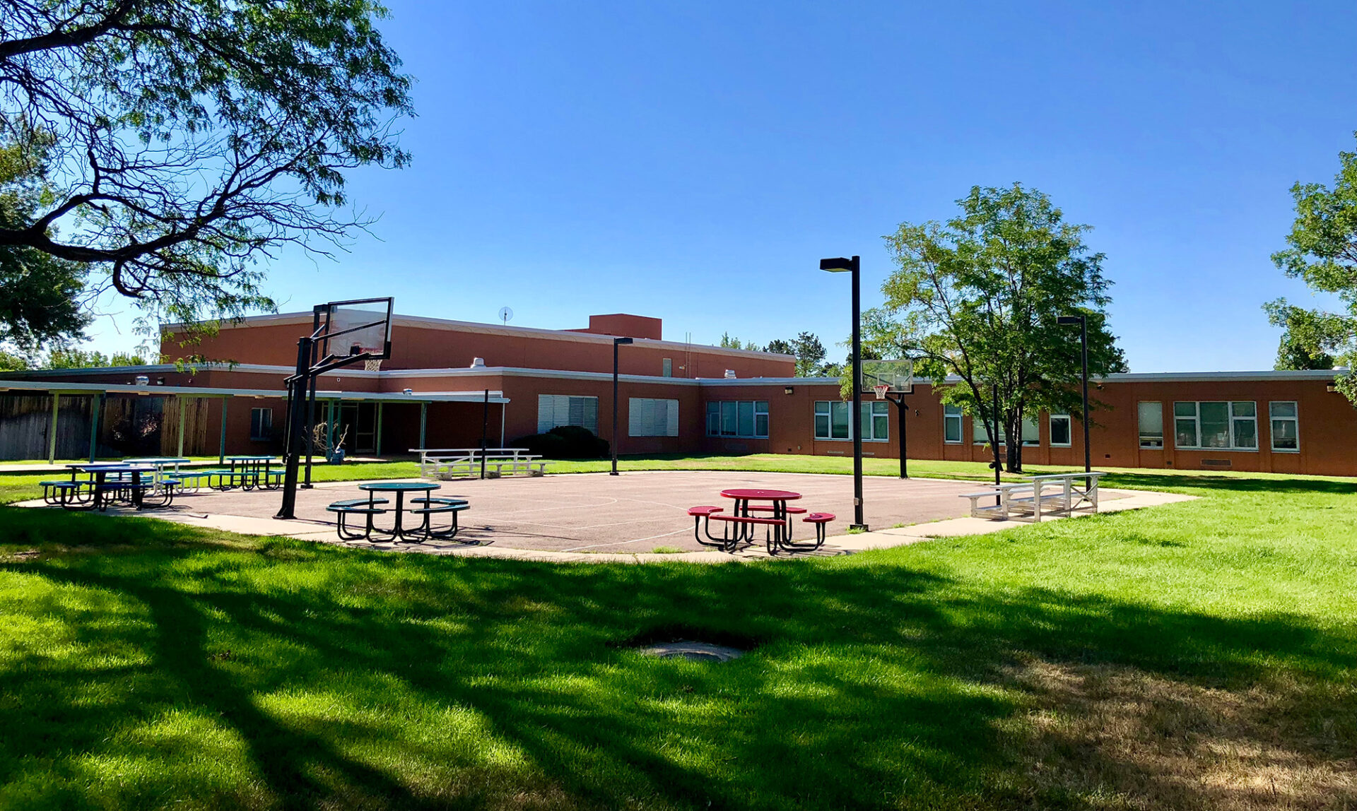 A peaceful outdoor recreational area with a basketball court, picnic tables painted red and blue, and green grass. Trees provide shade, and a single-story building with brick walls and large windows is visible in the background under a bright blue sky.