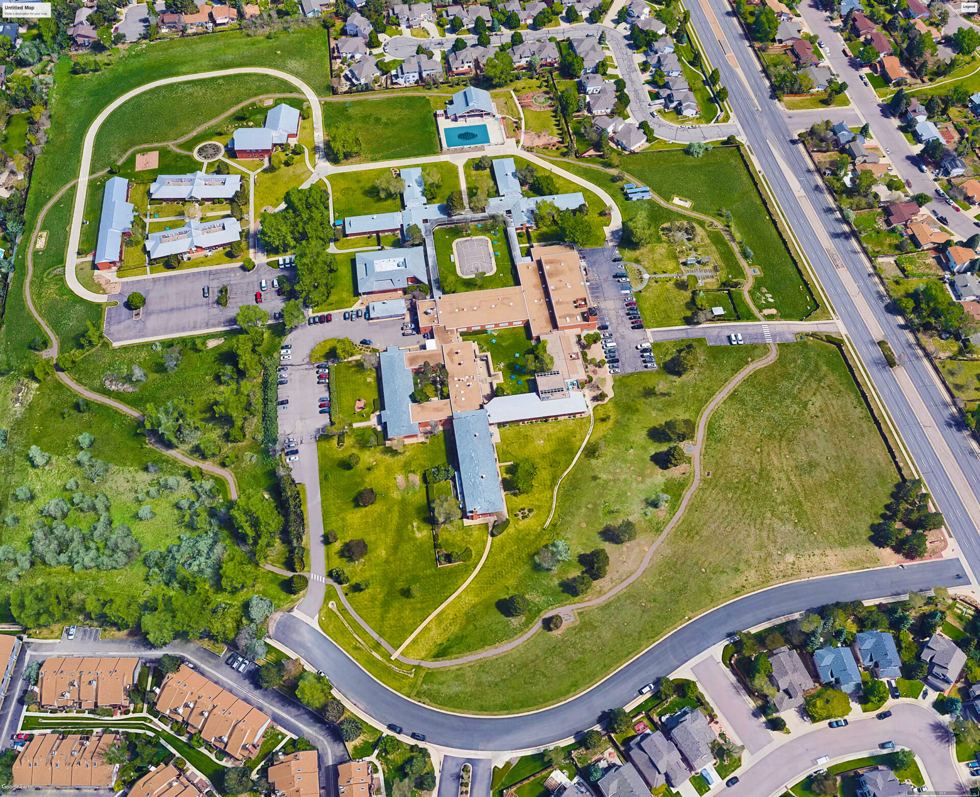 Aerial view of a large property with multiple buildings and green spaces, surrounded by residential neighborhoods. The site features pathways, parking lots, open grassy areas, and a distinct cluster of buildings, some with red roofs. Roads and sidewalks border the perimeter of the property.