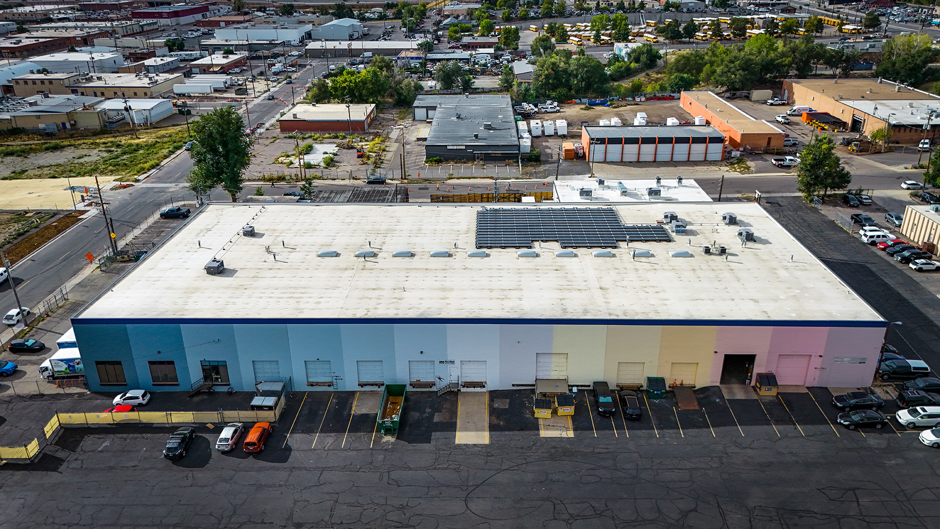 Aerial view of a large industrial building with a white roof, lined with colorful exterior walls transitioning from blue to pink. Solar panels are visible on part of the roof, and several cars are parked along the front. The surrounding area includes streets, trees, and industrial buildings.