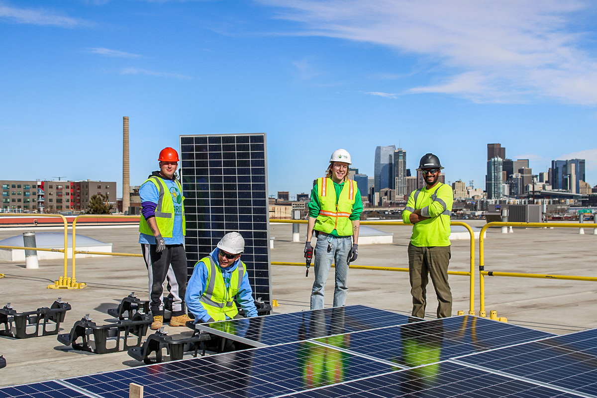 A group of four construction workers wearing bright safety vests, helmets, and gloves, working with solar panels on a rooftop. Behind them is a city skyline with tall buildings under a clear blue sky.