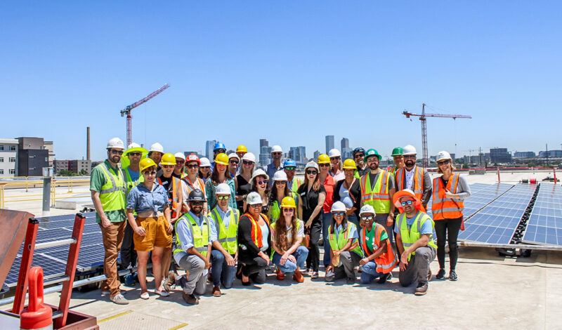 A large group of people, including construction workers and visitors, posing for a photo on a rooftop solar installation site. They are wearing hard hats and safety vests, and solar panels are spread across the roof. The city skyline and cranes are visible in the distance under a bright blue sky.