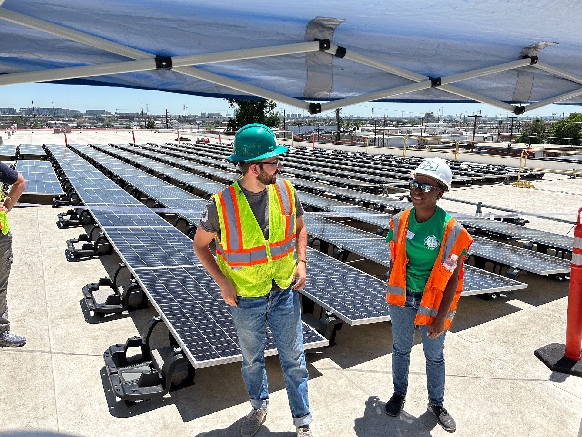 Two workers in bright safety vests and helmets standing under a shaded canopy on a rooftop surrounded by rows of installed solar panels. They appear to be discussing the project, with one smiling.