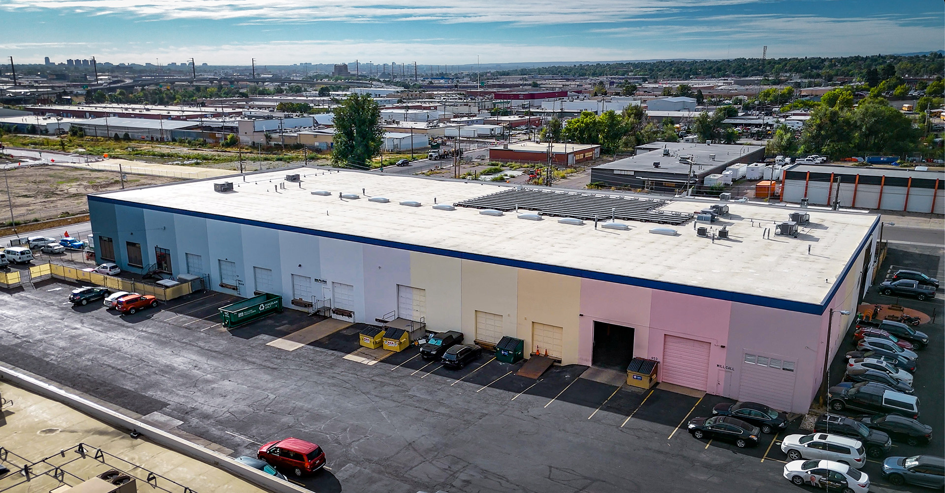 A ground-level perspective of the industrial building with rainbow-colored walls, a white roof, and solar panels on top. Several cars and dumpsters are parked in front, with nearby industrial buildings and streets visible in the background.