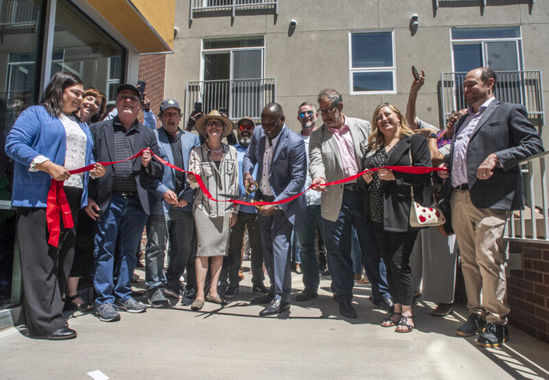 A group of people, including developers, city officials, and community leaders, participate in a ribbon-cutting ceremony outside a newly constructed apartment building during the grand opening of South Platte Crossing Apartments in Commerce City, Colorado.